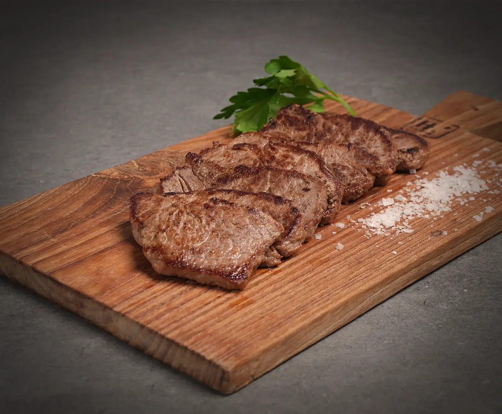 Sechs gebratene Fleischscheiben, vermutlich Rinderfilet oder Steak, liegen diagonal auf einem Holzschneidebrett. Daneben grobes Meersalz und ein Petersilienzweig auf dunklem Hintergrund.