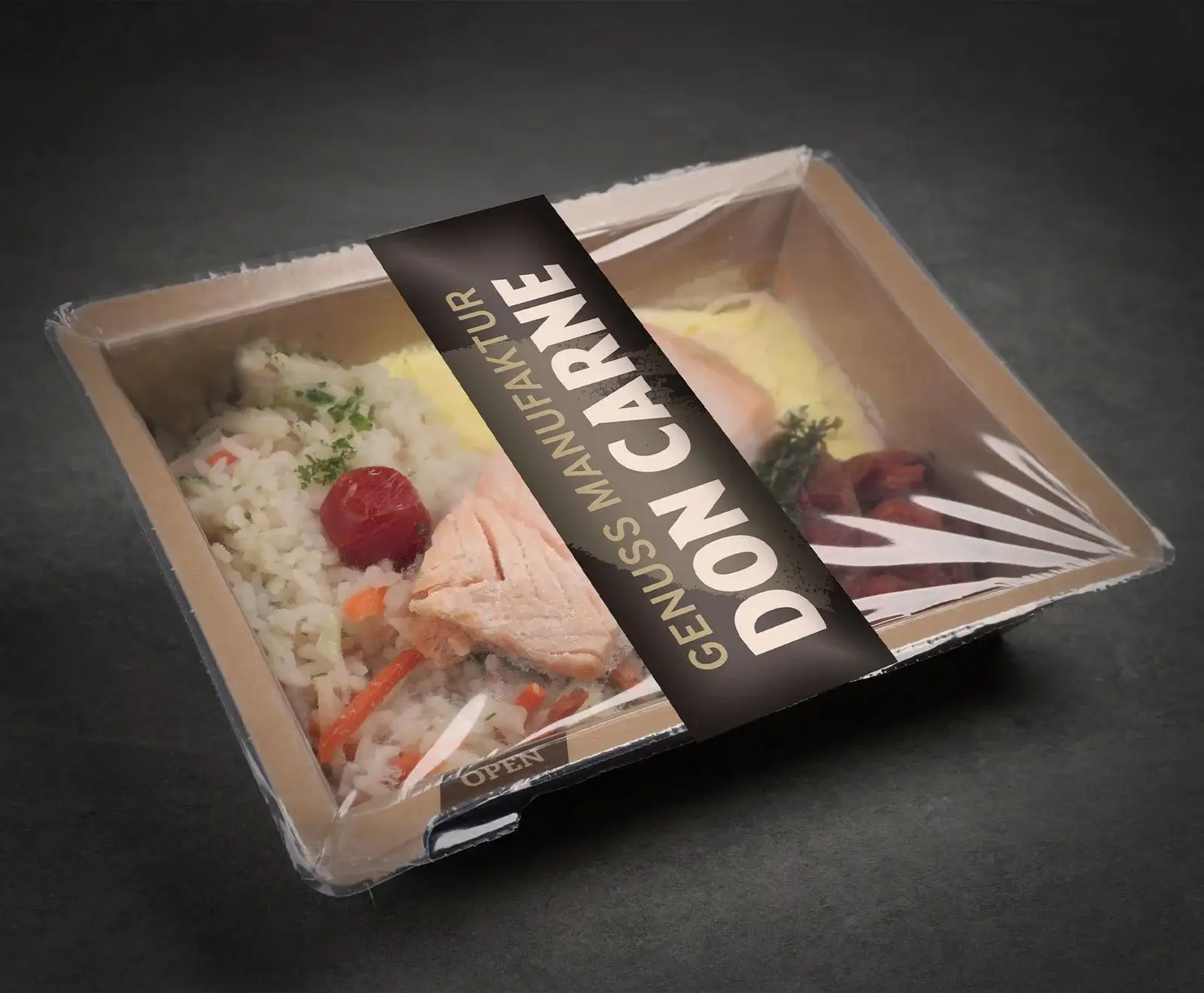 Eine Fertigmahlzeit in einer rechteckigen, hellbraunen Kartonschale mit transparentem Deckel, schräg von oben auf dunklem Hintergrund. Die Mahlzeit enthält Reis mit Gemüse, Lachs, eine Kirschtomate und weitere Beilagen. Ein schwarzes Etikett mit "GENUS