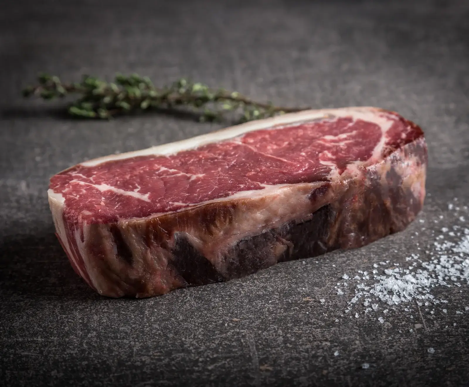 Nahaufnahme eines rohen, marmorierten Rindersteaks mit Fettdeckel auf dunklem Stein, begleitet von einem Thymianzweig und grobem Meersalz.