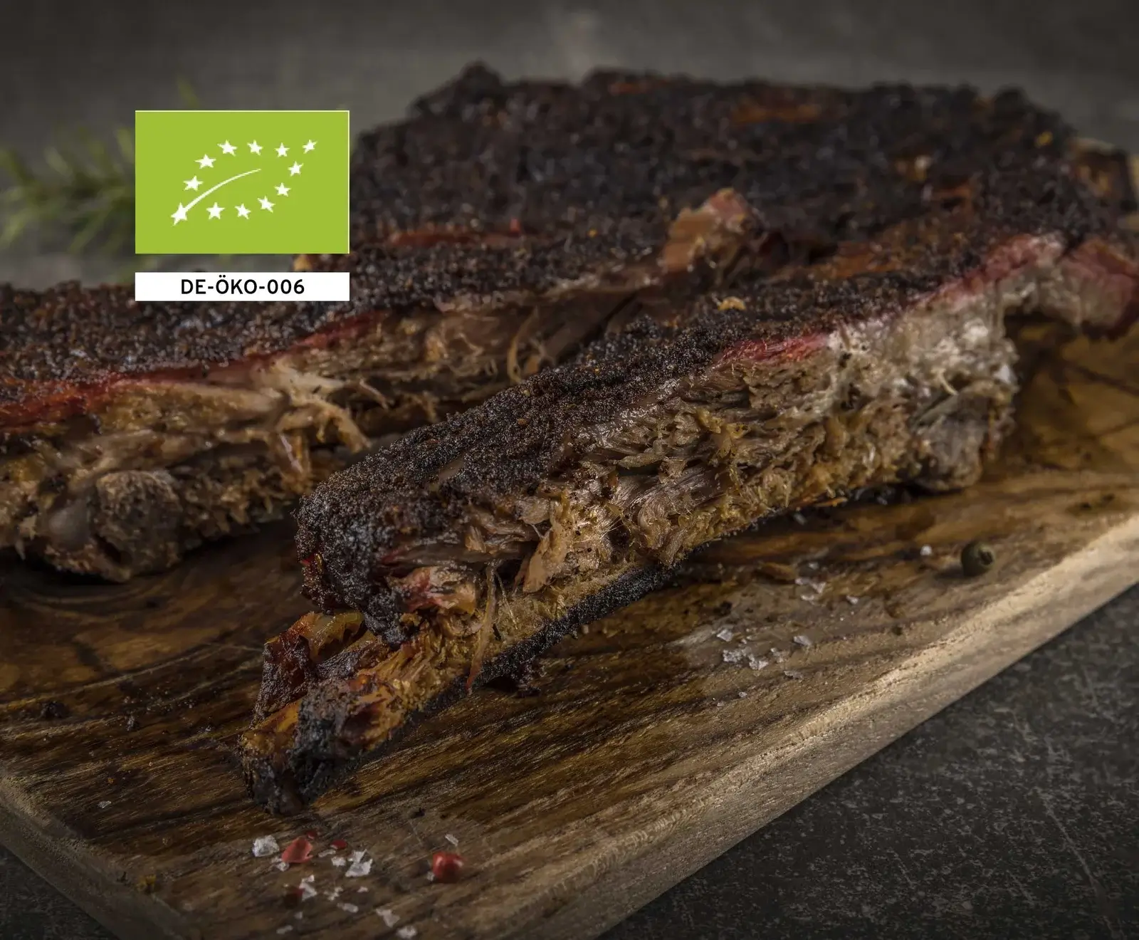 Nahaufnahme von zwei Stücken langsam gegartem, geräuchertem Fleisch mit dunkler Kruste und zartem Inneren auf einem Holzbrett, bestreut mit Salz und Pfeffer. Oben links das EU-Bio-Logo DE-ÖKO-006.