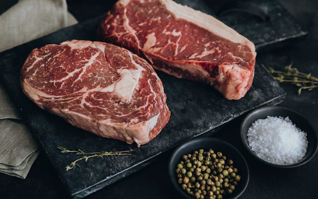Zwei rohe, marmorierte Steaks liegen auf einer dunklen Schieferplatte. Daneben stehen zwei kleine schwarze Schalen mit grobem Meersalz und grünen Pfefferkörnern. Einige Thymianzweige sind verstreut.
