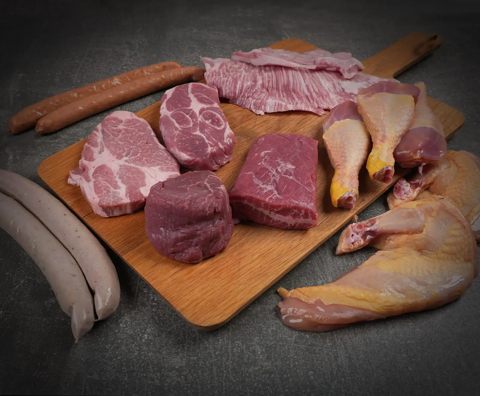 Eine vielfältige Auswahl an rohem Fleisch und Würsten auf einem Holzbrett und dunklem Hintergrund, darunter Schweinenackensteaks, Rindersteaks, Bauchspeck, Hähnchenkeulen, braune und weiße Würste.