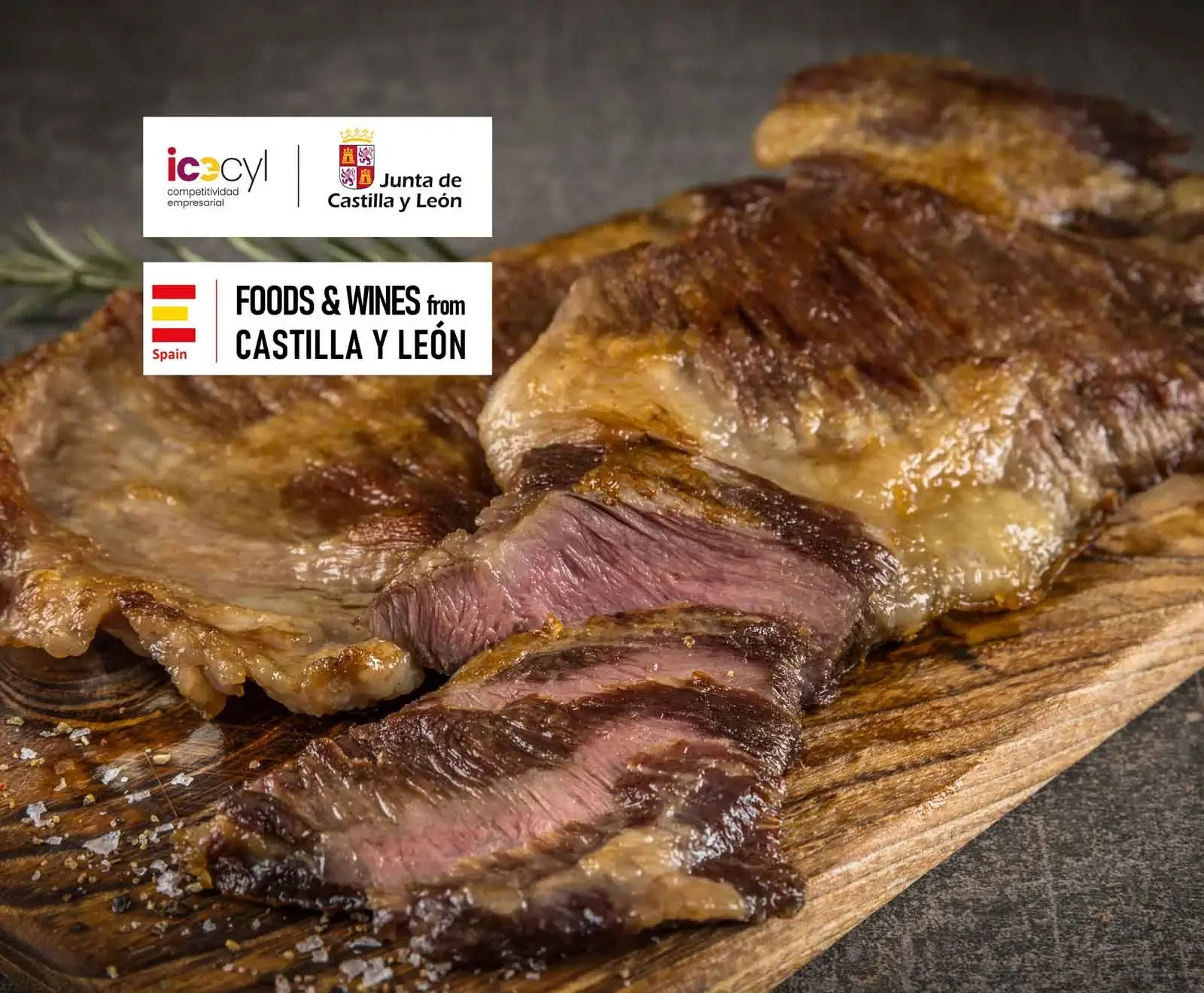 Ein gegrilltes Steak, teilweise aufgeschnitten mit rosa Kern, liegt auf einem Holzschneidebrett, bestreut mit grobem Salz. Im Hintergrund ein Rosmarinzweig und Logos von Castilla y León.