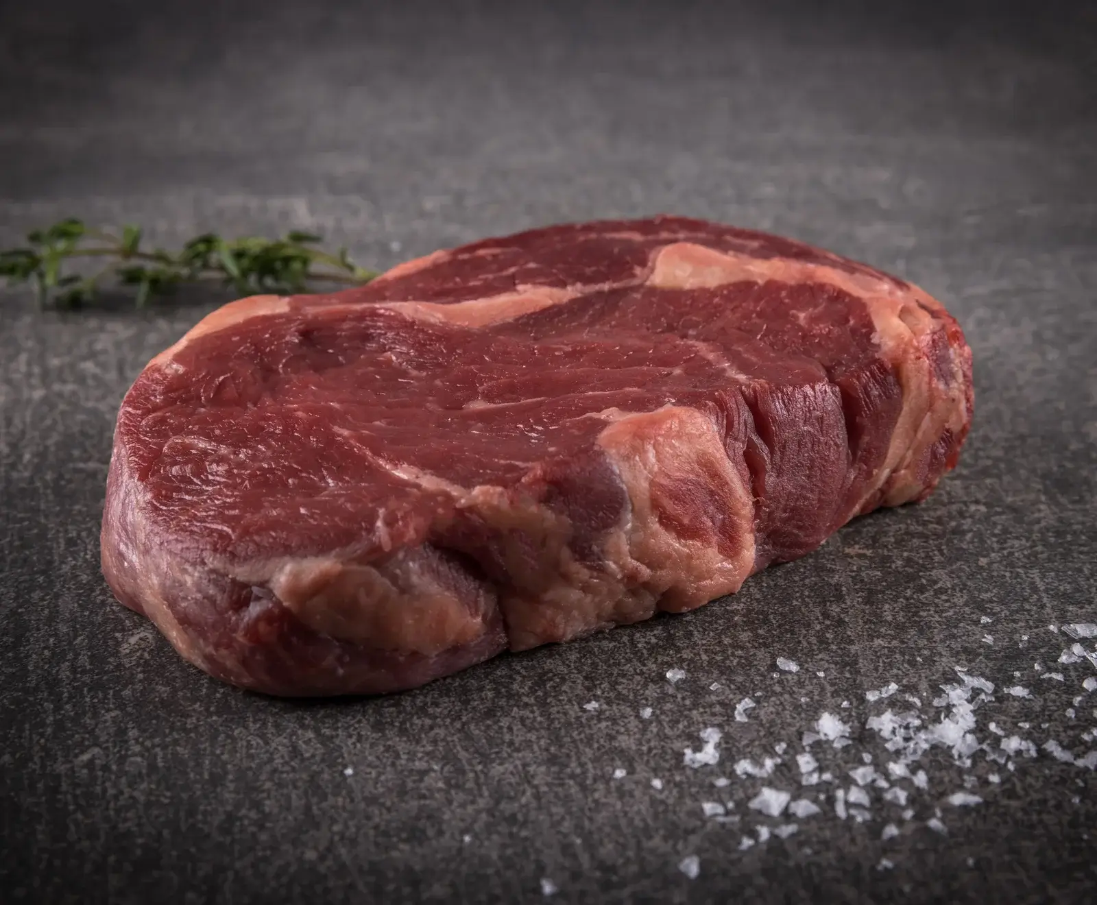 Ein rohes, marmoriertes Rindersteak liegt auf einer dunklen Steinoberfläche. Im Hintergrund ist ein Thymianzweig, im Vordergrund grobes Meersalz verstreut.