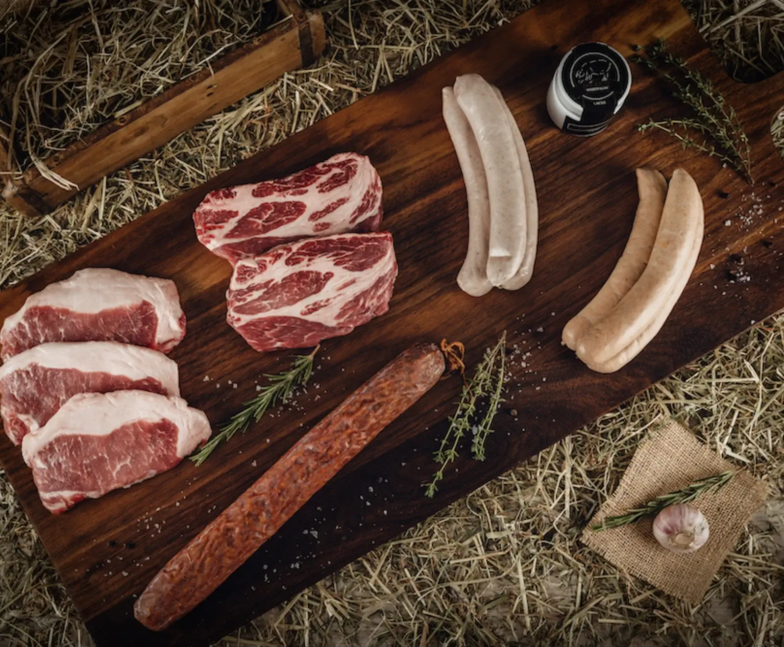Rustikale Anordnung von rohem Fleisch und Wurstwaren auf einem dunklen Holzbrett, umgeben von Heu. Zu sehen sind Schweinekoteletts, marmorierte Nackensteaks, helle und dunkle Bratwürste sowie eine Rohwurst, garniert mit Kräutern, Salz und Knoblauch.