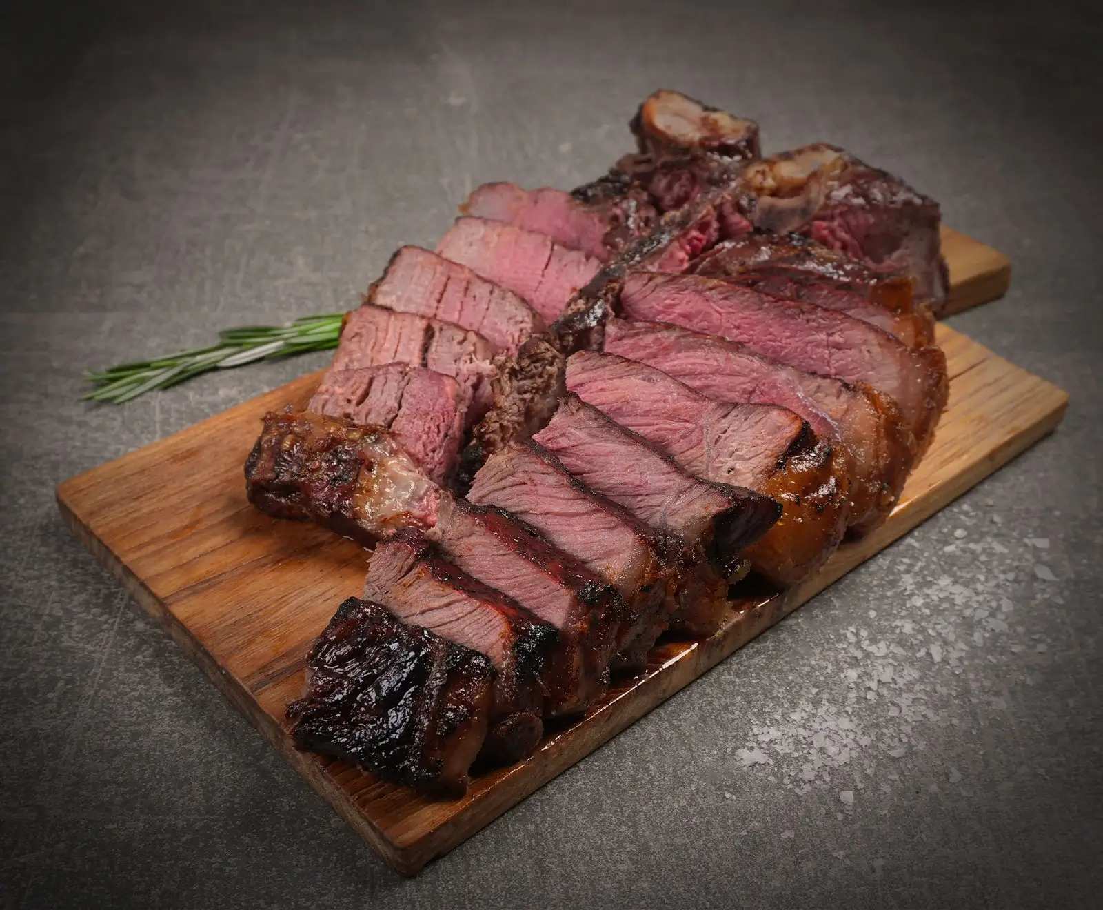 Ein saftiges, rosa gebratenes Steak, in dicke Scheiben geschnitten, liegt auf einem Holzbrett. Ein Rosmarinzweig ist daneben platziert. Der Hintergrund ist eine dunkle, strukturierte Oberfläche.