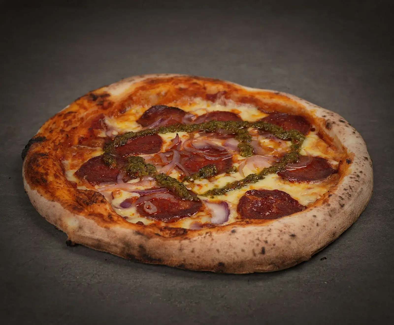 Eine frisch gebackene Pizza mit knusprigem Rand, belegt mit Salami, roten Zwiebeln, geschmolzenem Käse und einem Pesto-Drizzle, präsentiert auf dunklem Hintergrund.