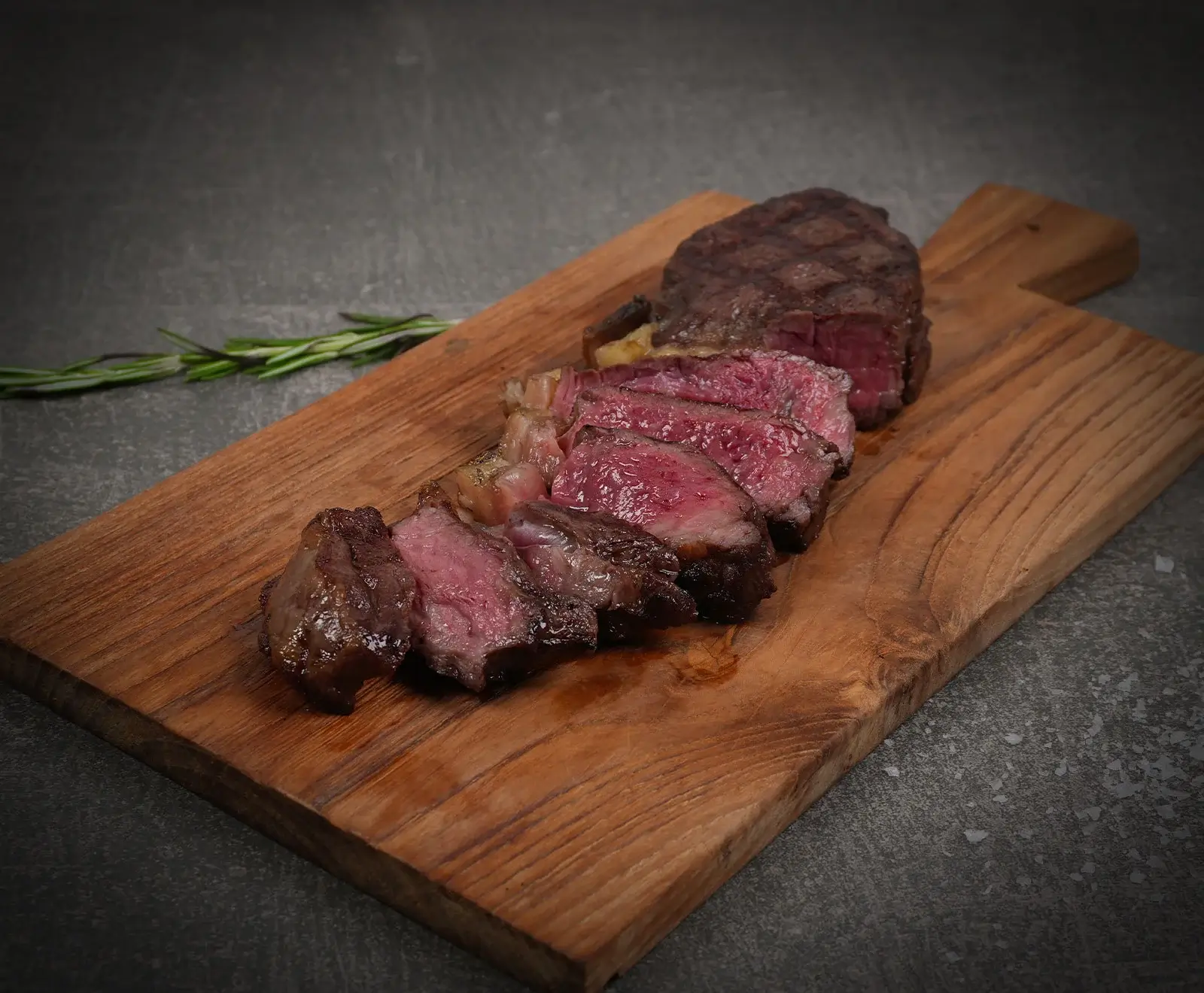 Ein perfekt gegartes, in Scheiben geschnittenes Steak, medium-rare, liegt auf einem rustikalen Holzschneidebrett. Im Hintergrund sind eine dunkle Oberfläche und ein Rosmarinzweig zu sehen.