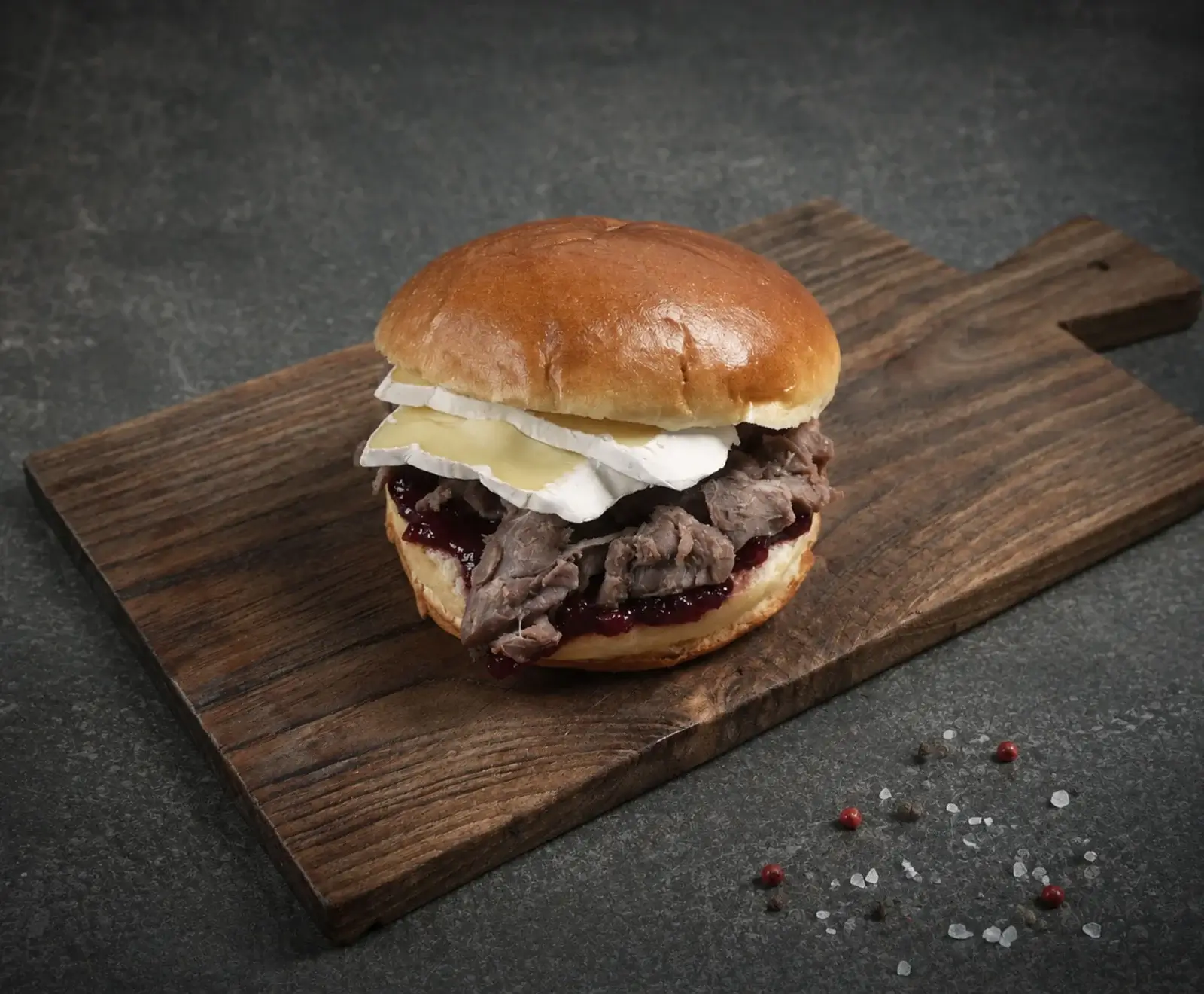Ein Gourmet-Burger mit goldbraunem Brioche-Brötchen, gezupftem Fleisch, dunkler Preiselbeersauce und mehreren Scheiben Brie-Käse, serviert auf einem rustikalen Holzbrett. Im Hintergrund eine dunkle Oberfläche mit verstreuten rosa Pfefferkörnern und gr