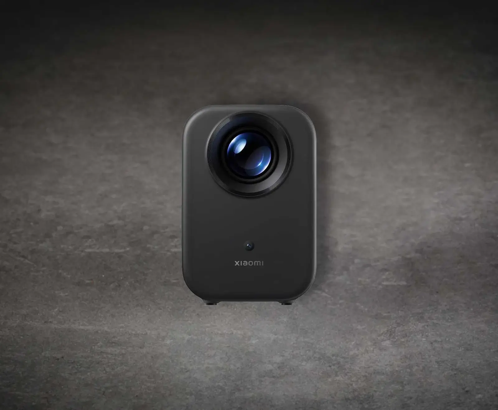 Ein schlanker, dunkelgrauer Xiaomi Projektor mit abgerundeten Ecken steht auf einer grauen Betonoberfläche. Die große, runde Linse oben reflektiert blaues Licht, darunter ist das Xiaomi-Logo zu sehen.