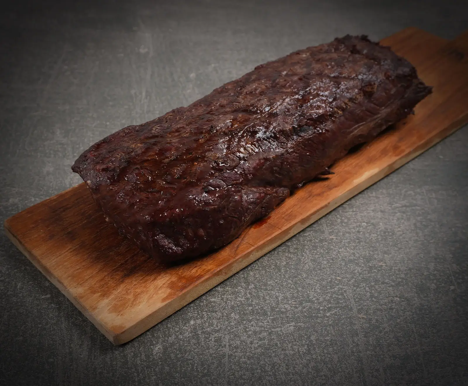 Ein großes, dunkelbraun gebratenes Stück Fleisch, glänzend und saftig aussehend, liegt auf einem hellbraunen Holzschneidebrett. Der Hintergrund ist eine dunkle, graue Oberfläche.