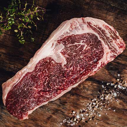 Ein rohes, stark marmoriertes Rindersteak liegt auf einem dunklen Holzbrett. Links oben sind frische Kräuter, rechts unten grobes Salz und Pfeffer verstreut.