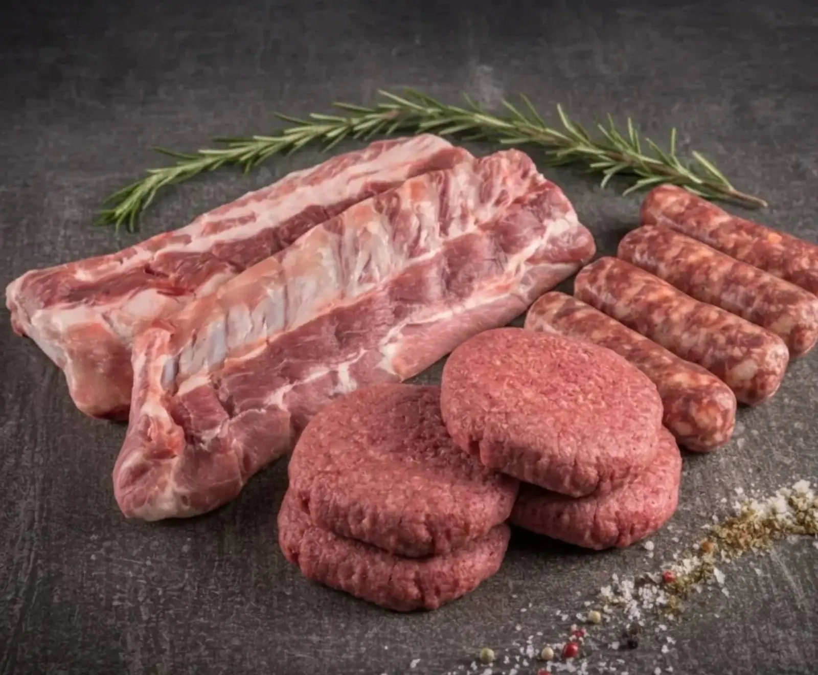 Rohe Schweinerippchen, Burger-Patties und Würstchen liegen auf einer dunklen Oberfläche, garniert mit Rosmarin und Gewürzen.
