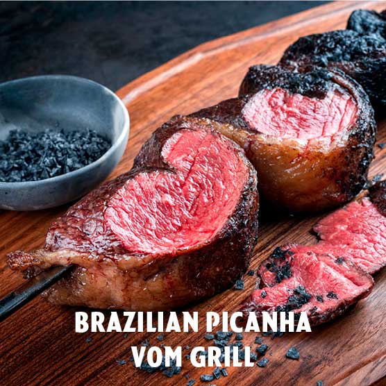Nahaufnahme von gegrilltem brasilianischem Picanha-Steak, medium-rare, auf einem Spieß und geschnitten auf einem Holzbrett, garniert mit schwarzem Salz. Eine Schale mit Salz steht daneben.