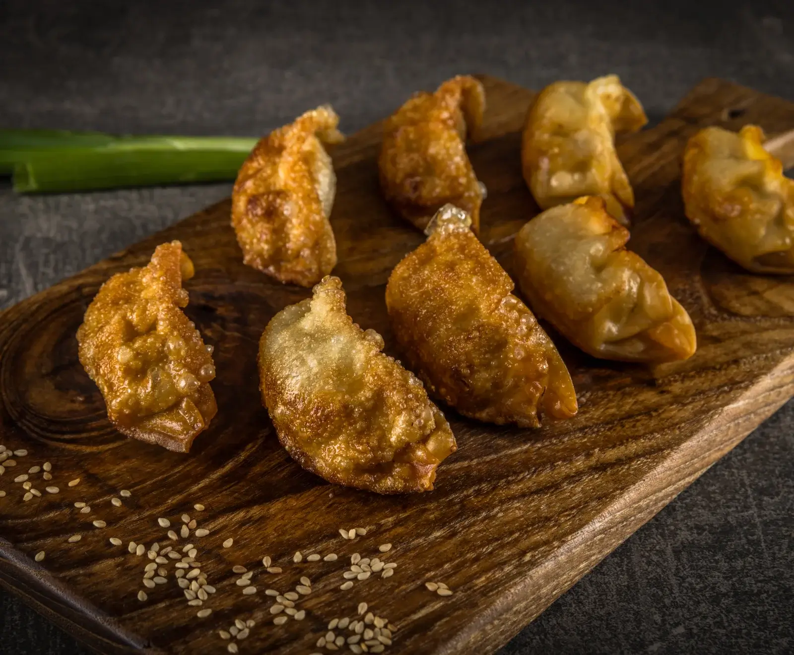 Mehrere goldbraun gebratene Teigtaschen (Gyoza) liegen auf einem dunklen Holzbrett, bestreut mit Sesamsamen. Im Hintergrund ist eine grüne Lauchzwiebel auf dunklem Untergrund zu sehen.
