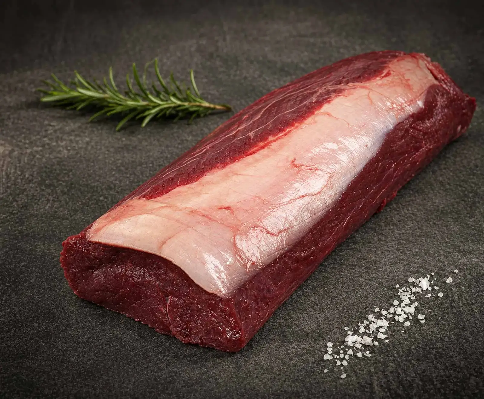 Ein großes, rohes Stück dunkelrotes Fleisch, vermutlich ein Filet oder Lende, mit einer weißen Fettschicht. Ein Rosmarinzweig und grobes Meersalz liegen auf dunklem Untergrund.