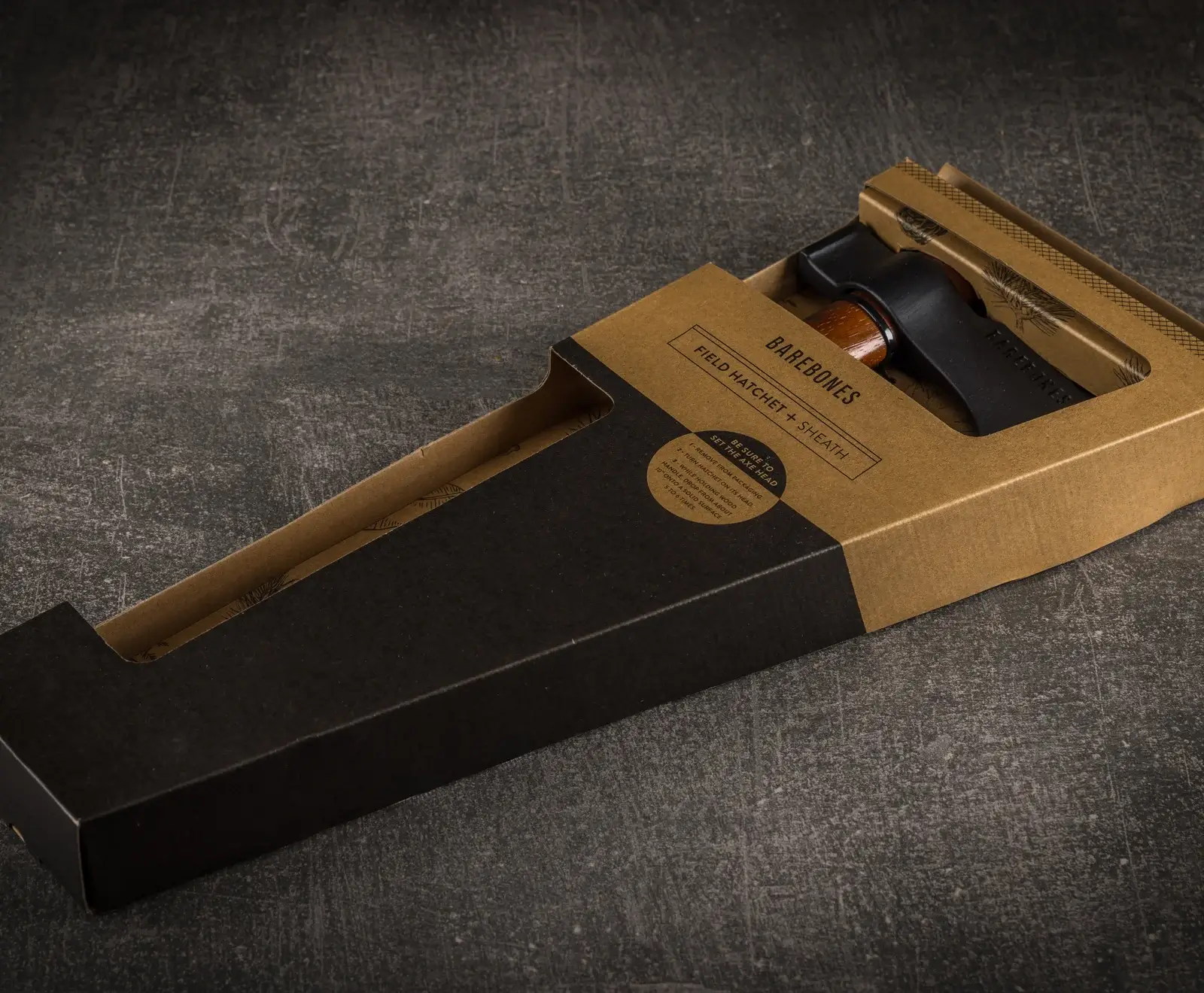 Ein Barebones Feldbeil mit Scheide in seiner braun-schwarzen Kartonverpackung, auf einem dunklen, strukturierten Untergrund. Der Holzgriff des Beils und die schwarze Scheide sind sichtbar.