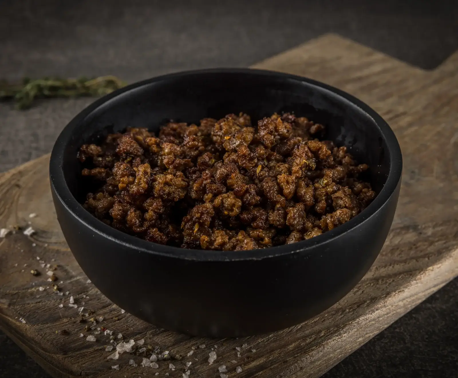 Eine schwarze Schale gefüllt mit gebratenem, braunem Hackfleisch oder veganem Hack, auf einem rustikalen Holzbrett mit verstreutem Salz und Pfeffer. Im Hintergrund sind Kräuter zu sehen.