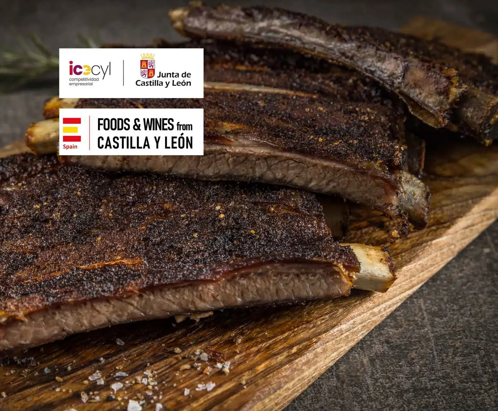 Dunkel gewürzte Rippchen auf einem Holzbrett mit Salzkristallen. Links oben: Logos der Junta de Castilla y León und "Foods & Wines from Castilla y León, Spain".