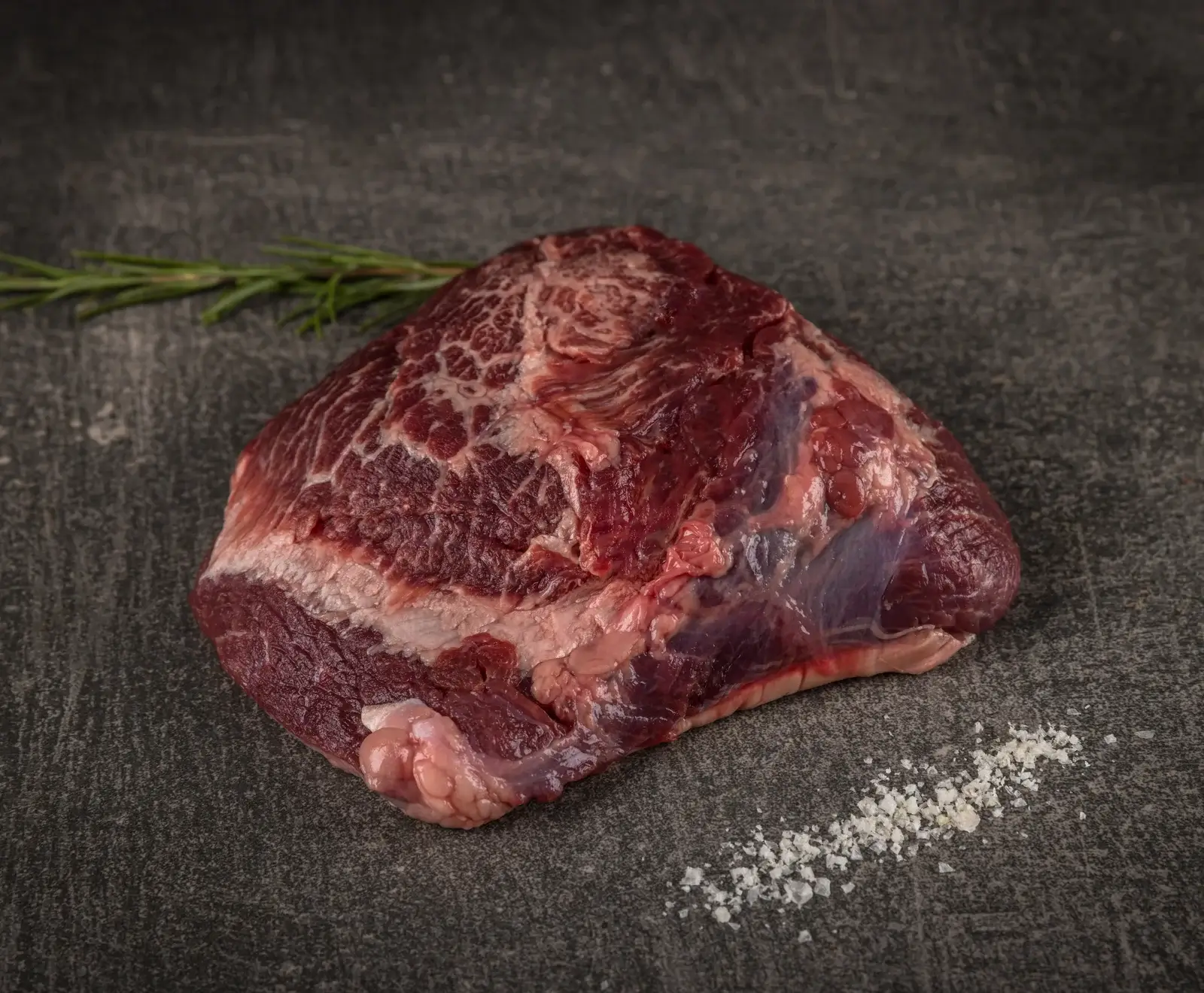 Ein rohes Stück Rindfleisch mit deutlicher Marmorierung liegt auf einer dunklen Steinplatte. Daneben befinden sich ein Rosmarinzweig und eine Prise grobes Salz.