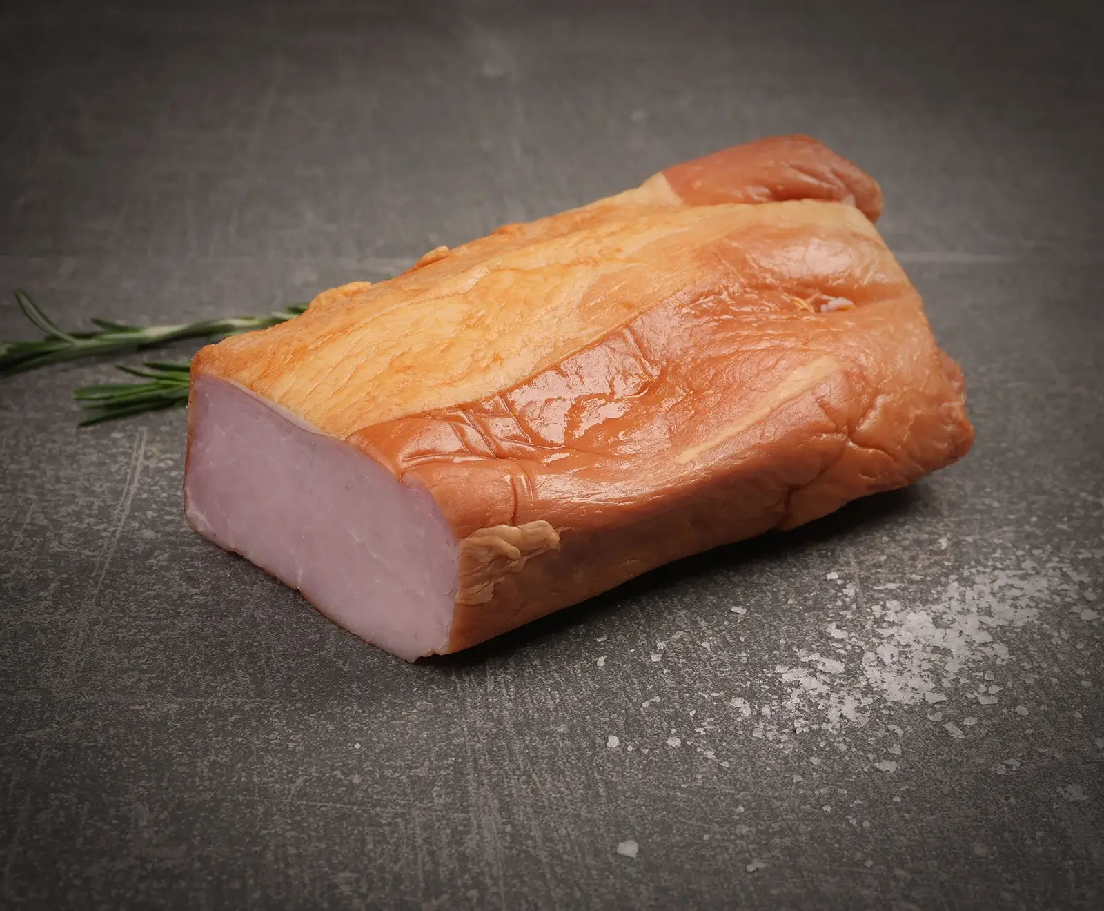Ein Stück geräucherter Schweinelachs mit goldbrauner Kruste und rosa Schnittfläche liegt auf einer dunklen Steinplatte. Daneben Rosmarin und grobes Salz.
