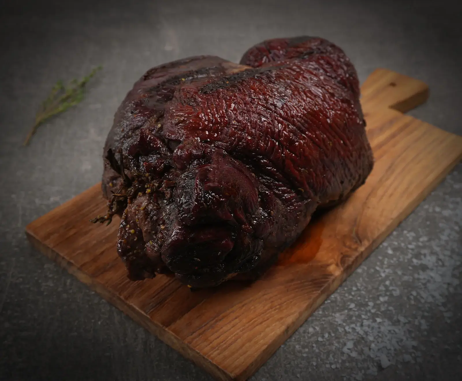 Dunkelbraun glänzender, gebratener oder geräucherter Fleischbraten auf einem rustikalen Holzbrett, mit Kräutern gewürzt, vor dunklem Hintergrund mit Rosmarinzweig.
