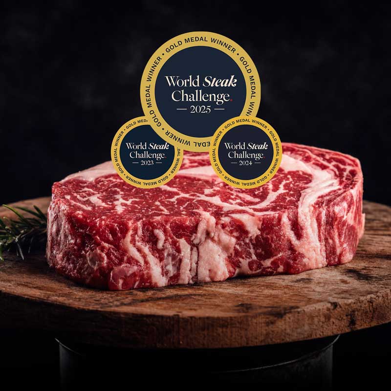 Ein rohes, stark marmoriertes Ribeye-Steak liegt auf einem Holzbrett mit Rosmarin. Darüber schweben drei goldene "World Steak Challenge Gold Medal Winner"-Abzeichen für die Jahre 2023, 2024 und 2025.