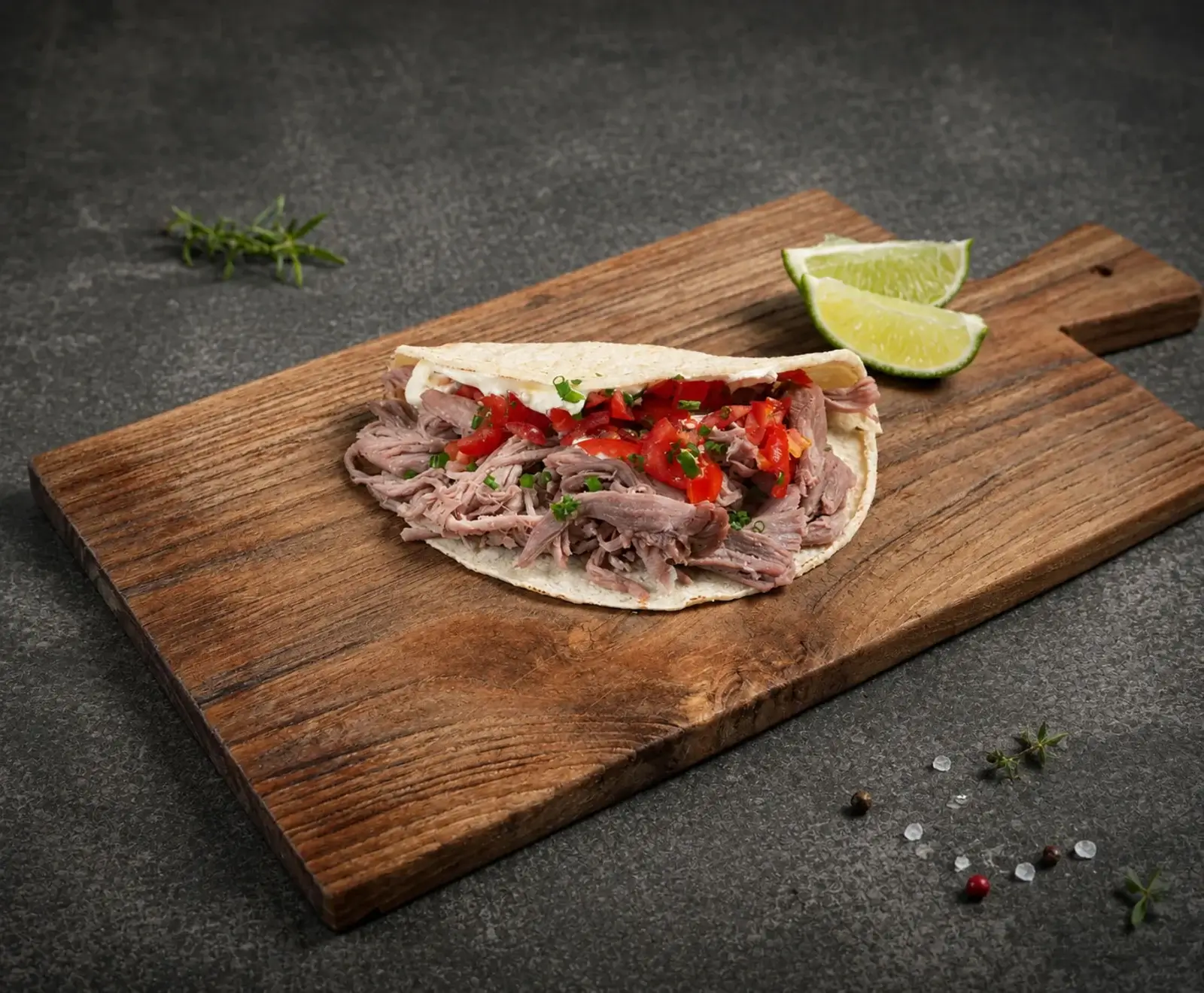 Ein offener Taco oder Wrap mit zerkleinertem Fleisch, gewürfelten Tomaten und Schnittlauch, serviert auf einem rustikalen Holzbrett. Daneben liegen zwei Limettenspalten und Gewürze auf dunklem Untergrund.