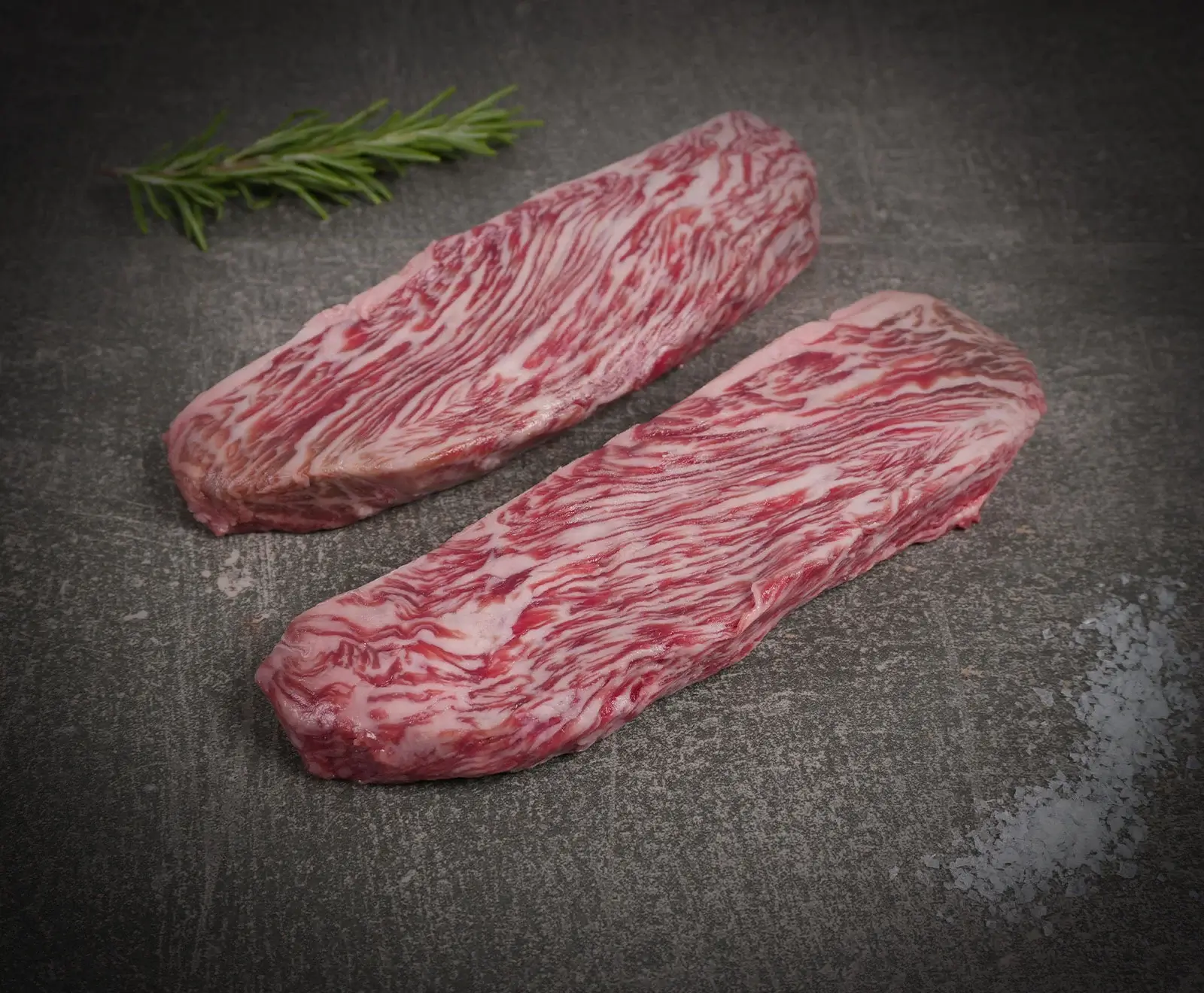 Zwei rohe, stark marmorierte Rindersteaks liegen auf einer dunklen Oberfläche. Links oben ein Rosmarinzweig, rechts unten grobes Meersalz. Das Fleisch zeigt eine intensive Fettzeichnung.