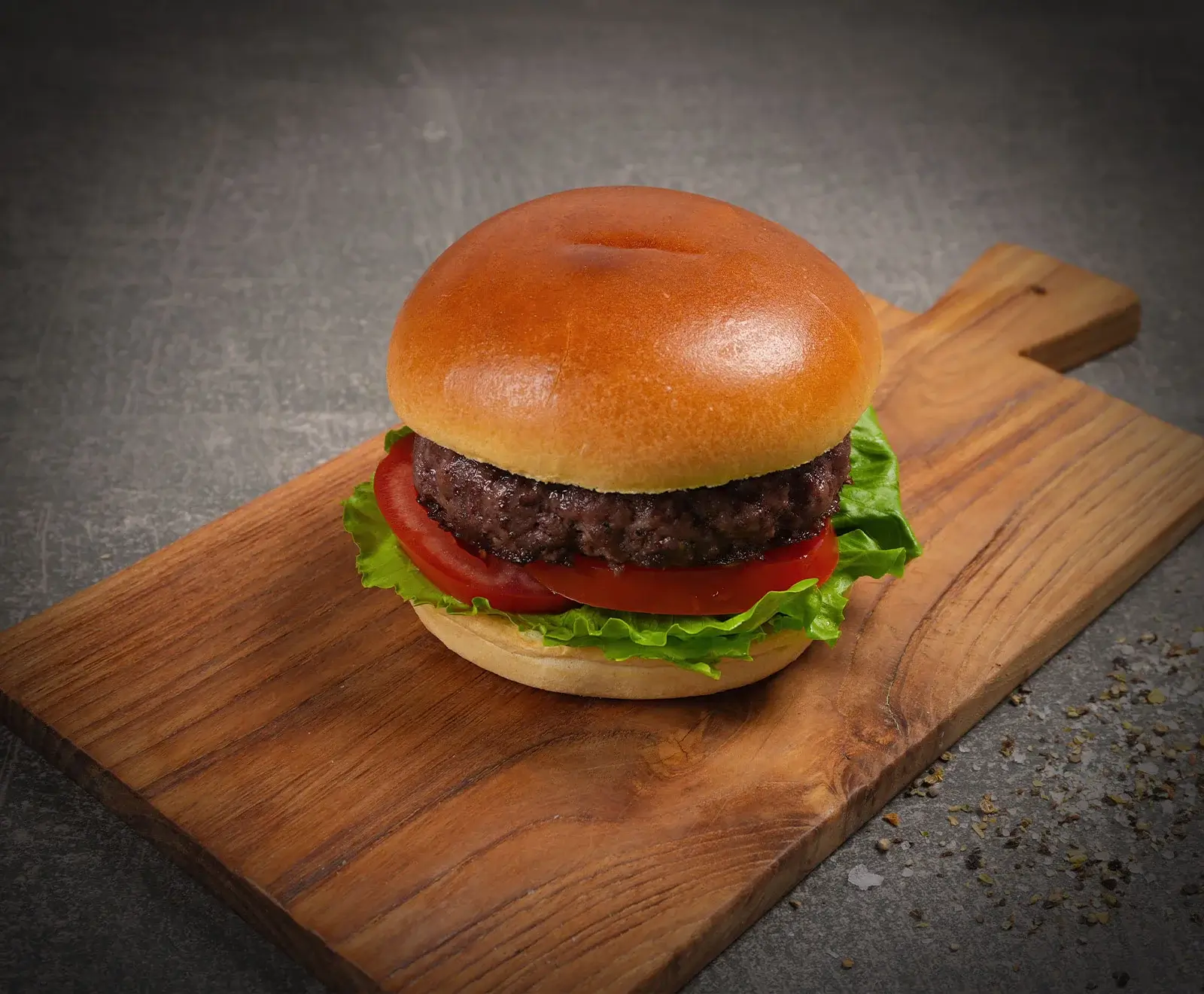 Ein klassischer Burger mit goldbraunem Brioche-Brötchen, saftigem Rindfleisch-Patty, frischem grünen Salat und roten Tomatenscheiben, serviert auf einem Holzschneidebrett vor dunklem Hintergrund.
