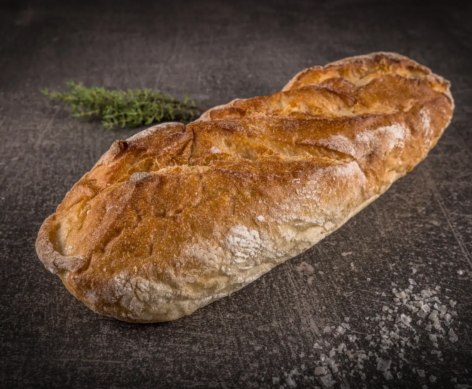 Ein goldbrauner, rustikaler Brotlaib mit bemehlter, aufgeschnittener Kruste liegt diagonal auf einer dunklen, strukturierten Oberfläche. Daneben liegen ein Thymianzweig und grobes Salz.