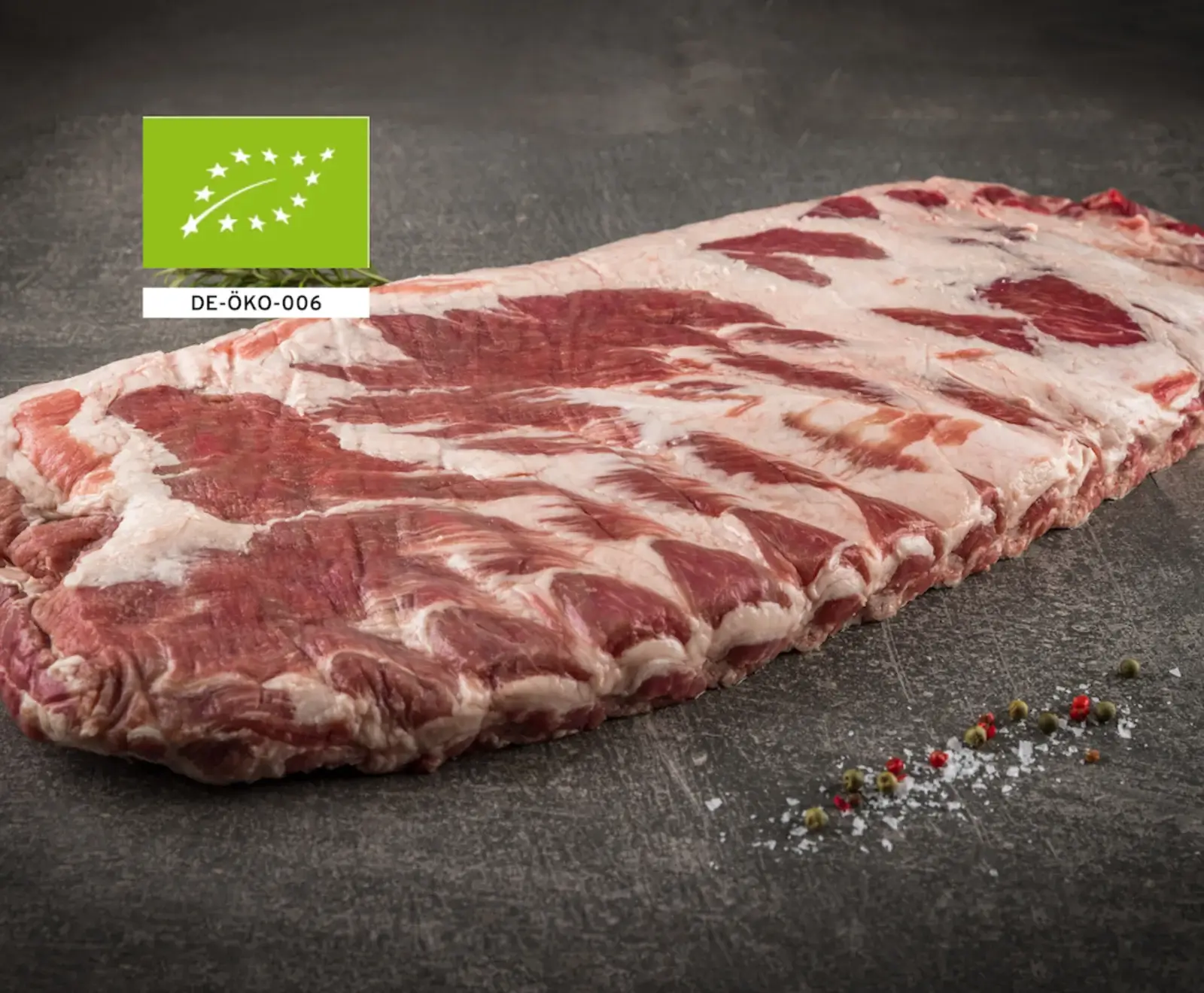 Rohe Schweinerippchen mit viel Fleisch und Fett auf dunklem Untergrund, daneben Salz und Pfeffer. Oben links ist das EU-Bio-Siegel mit DE-ÖKO-006 zu sehen.
