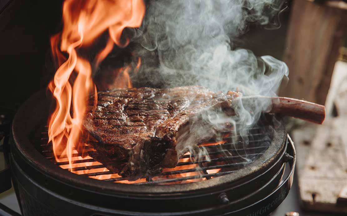Ein großes Tomahawk-Steak mit Knochen liegt auf einem Kamado-Grill. Helle Flammen schlagen links vom Fleisch hoch, während dichter Rauch aufsteigt. Das Steak hat deutliche Grillspuren.