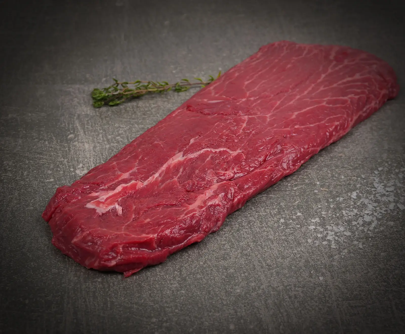 Ein rohes, flaches Stück rotes Rindfleisch (Flank Steak) liegt auf einer dunklen Steinoberfläche. Daneben ein Zweig frischer Thymian und grobes Salz.