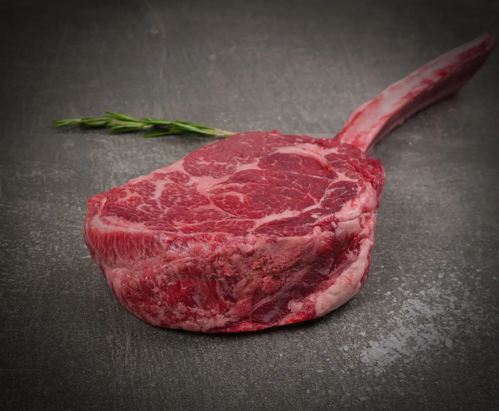 Ein rohes Tomahawk-Steak mit Knochen und deutlicher Marmorierung liegt auf einer dunklen, strukturierten Oberfläche. Im Hintergrund ist ein Rosmarinzweig zu sehen.