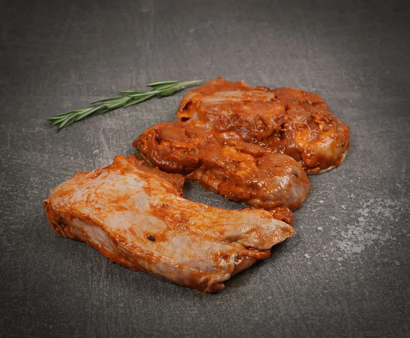 Zwei rohe, rot-orange marinierte Fleischstücke liegen auf einer dunklen Steinplatte. Ein Rosmarinzweig und grobe Salzkristalle sind im Hintergrund und rechts daneben zu sehen.