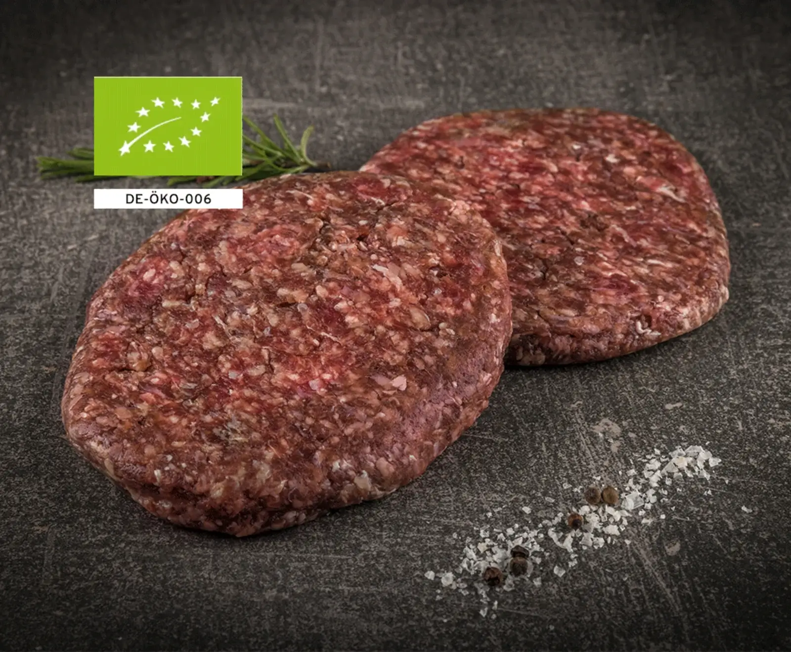 Zwei rohe Bio-Rinder-Burgerpatties auf dunklem Schiefer, garniert mit Rosmarin, grobem Salz und Pfeffer. Oben links ist das EU-Bio-Siegel mit DE-ÖKO-006 zu sehen.