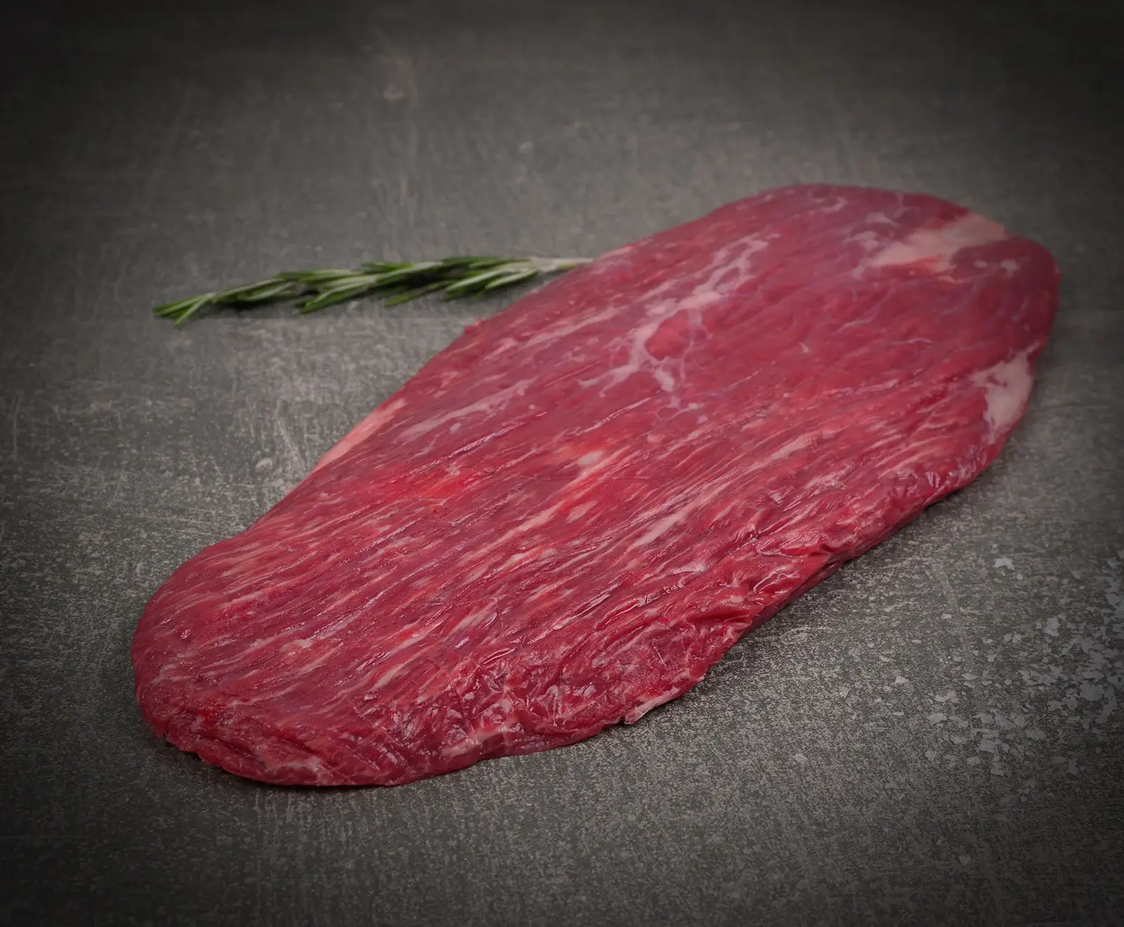Ein rohes, längliches Flanksteak mit sichtbarer Marmorierung liegt auf einer dunklen, strukturierten Oberfläche. Im Hintergrund ist ein Rosmarinzweig zu sehen.