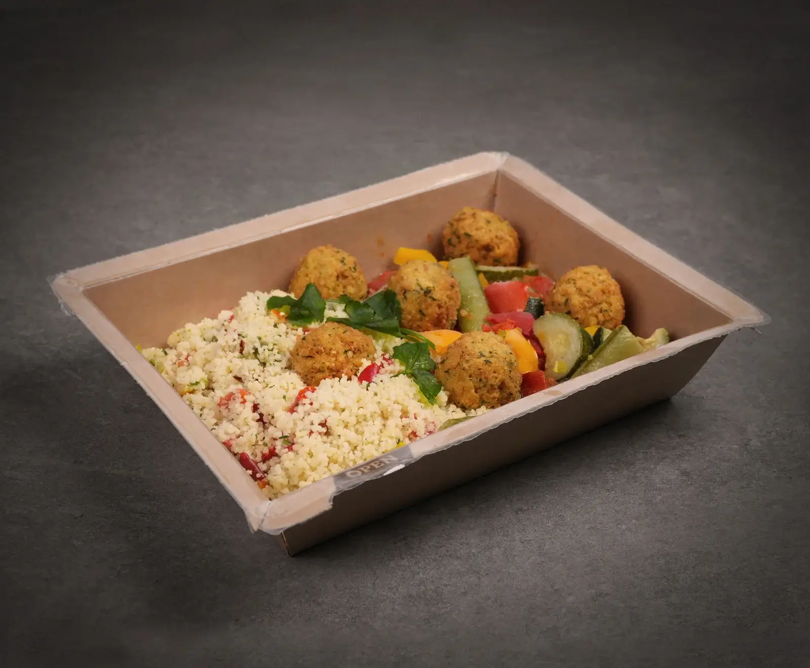 Eine rechteckige, braune Take-away-Schale mit Couscous, Falafelbällchen und gehacktem Gemüse wie Gurke, Tomate und Paprika, garniert mit frischen Kräutern, auf dunklem Hintergrund.