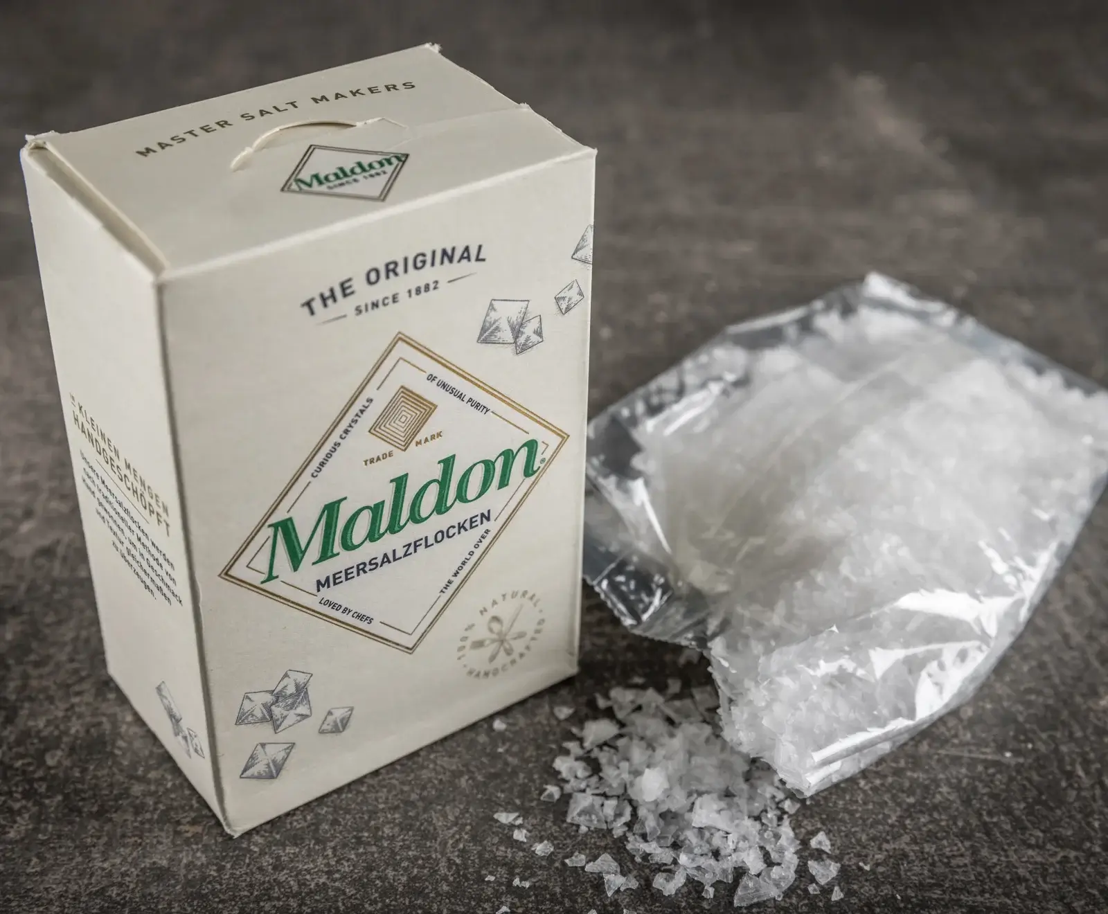 Eine beige Schachtel Maldon Meersalzflocken steht neben einem geöffneten Plastikbeutel mit weißen Salzflocken, die teilweise auf eine dunkle Oberfläche verschüttet sind. Die Schachtel zeigt das grüne Maldon-Logo und den Text "THE ORIGINAL SINCE 1882"