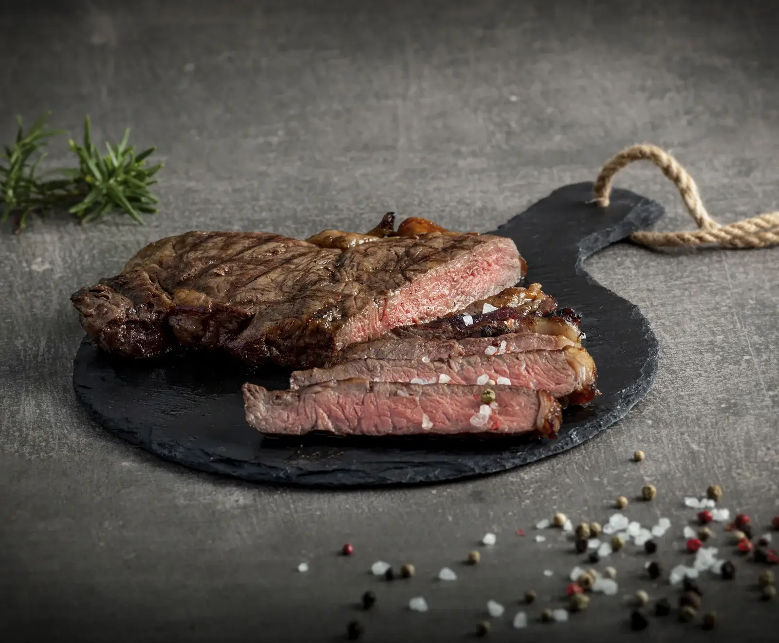 Ein perfekt gegrilltes, aufgeschnittenes Steak (medium-rare) auf einer schwarzen Schieferplatte, garniert mit Rosmarin. Grobes Meersalz und bunte Pfefferkörner liegen auf dem dunklen Untergrund.