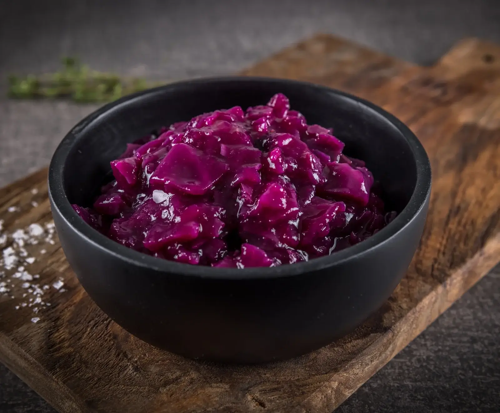 Eine schwarze Schale gefüllt mit leuchtend violettem, gekochtem Rotkohl, der auf einem rustikalen Holzbrett steht. Im Hintergrund sind unscharfe Kräuter zu sehen.