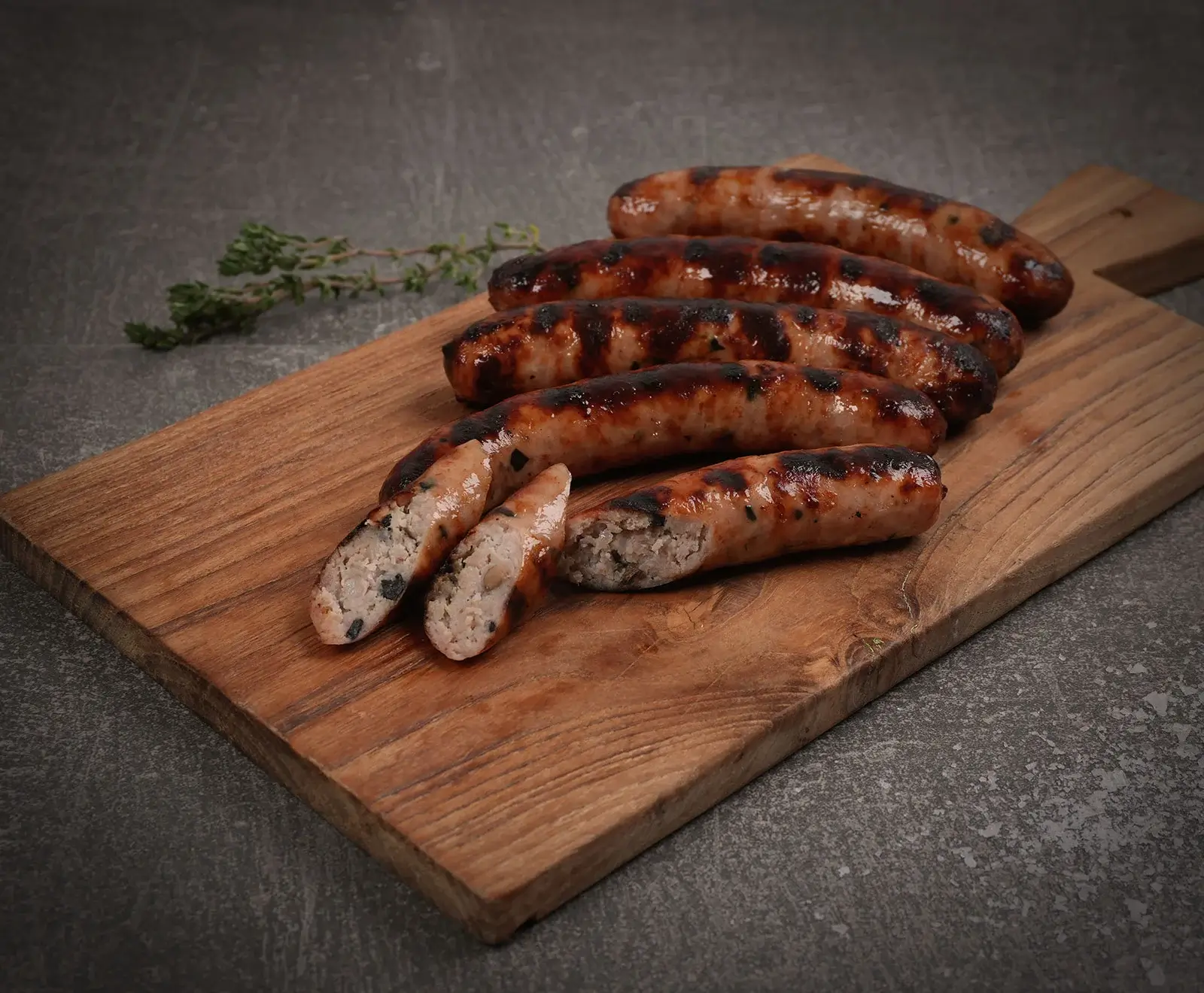 Fünf goldbraun gegrillte Würste auf einem Holzbrett, eine davon aufgeschnitten, um die helle Füllung mit dunklen Kräutern zu zeigen. Im Hintergrund liegen Thymianzweige.