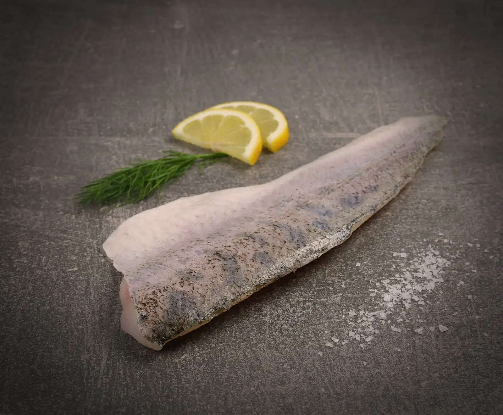 Ein rohes Fischfilet mit Haut liegt auf einer dunkelgrauen Oberfläche, daneben zwei Zitronenspalten, ein Dillzweig und grobes Meersalz.