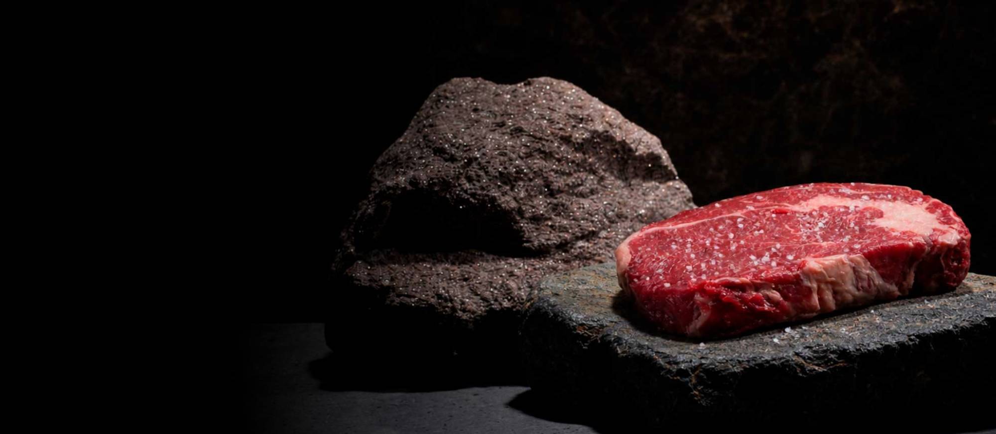 Ein rohes, mit grobem Salz bestreutes Steak liegt auf einem dunklen, flachen Stein. Im Hintergrund ist ein großer, dunkler, strukturierter Felsen zu sehen. Dunkle, dramatische Beleuchtung.