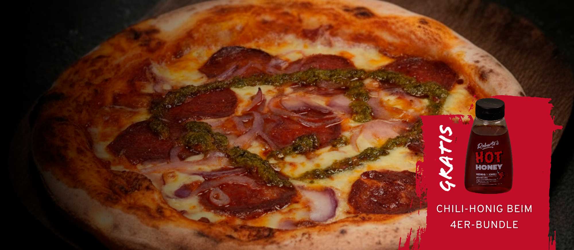 Eine knusprige Pizza mit Peperoni, roten Zwiebeln und grünem Pesto. Rechts unten ein rotes Feld mit einer Flasche Roberto's Hot Honey und dem Text "GRATIS CHILI-HONIG BEIM 4ER-BUNDLE".