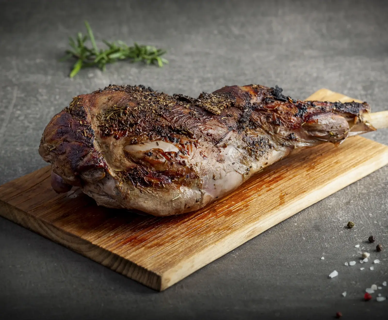 Eine knusprig gebratene, goldbraune Lammkeule, reichlich mit Kräutern gewürzt, liegt auf einem Holzbrett. Im Hintergrund ist ein Rosmarinzweig, im Vordergrund Pfefferkörner und grobes Salz.