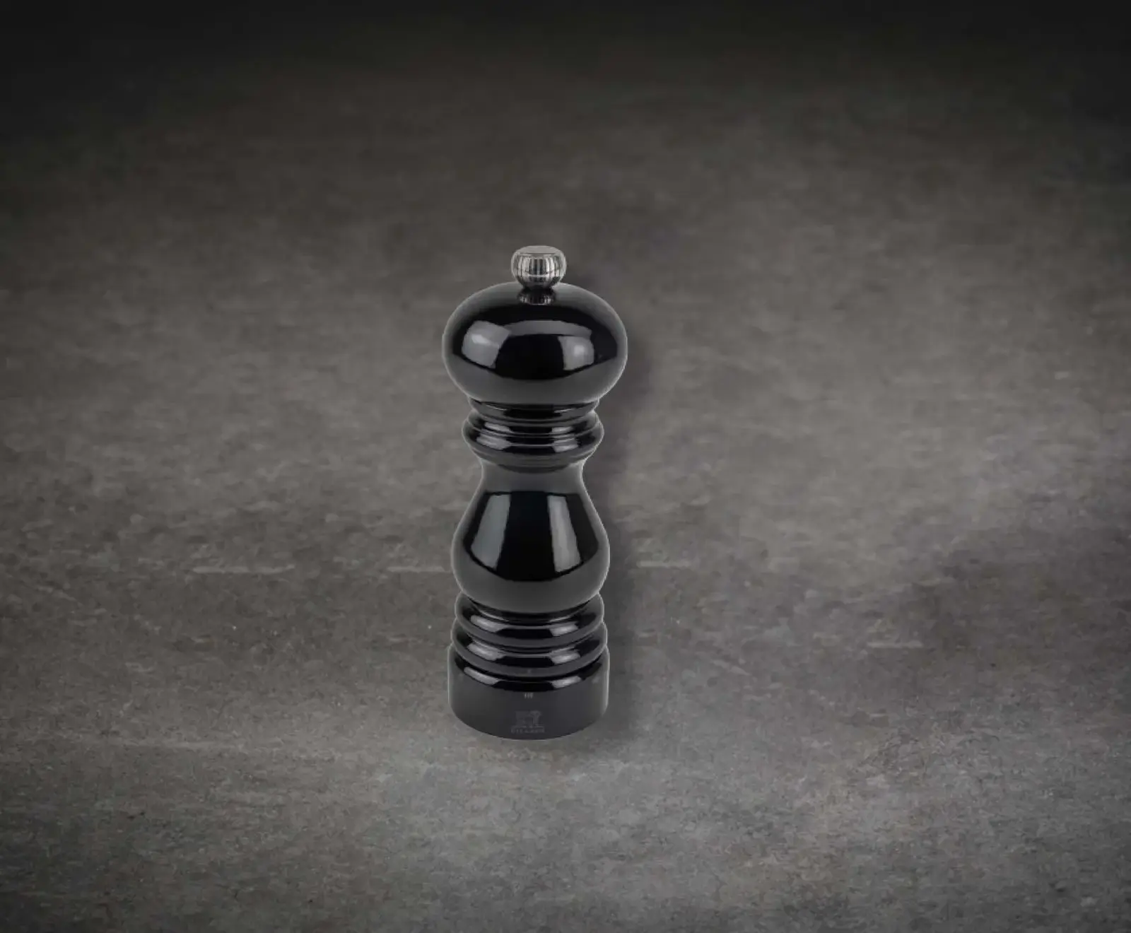 Eine glänzende, schwarze Pfeffermühle mit silbernem Knopf und Peugeot-Logo steht auf einem dunklen, strukturierten Hintergrund.