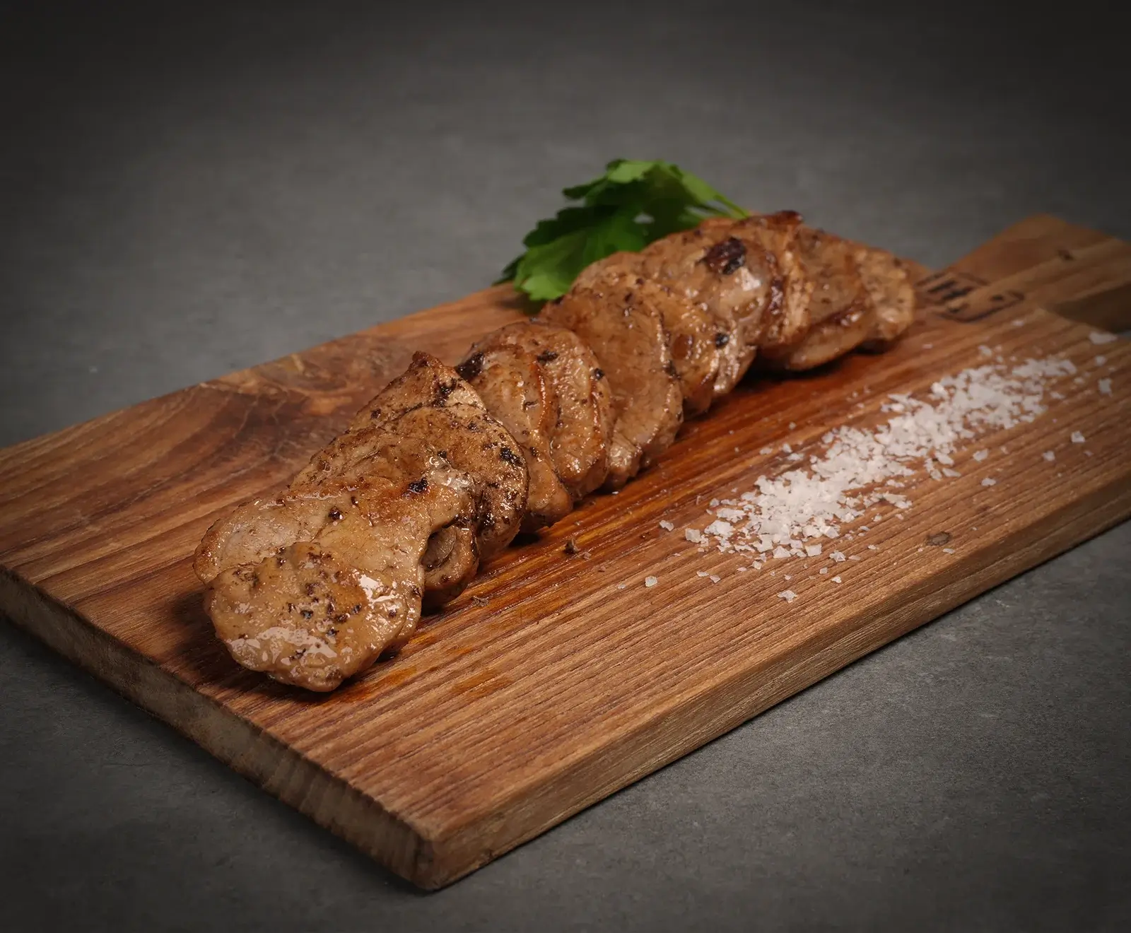Nahaufnahme von gebratenen, in Scheiben geschnittenen Fleischstücken auf einem Holzbrett, garniert mit einem Petersilienzweig und grobem Meersalz, vor dunklem Hintergrund.