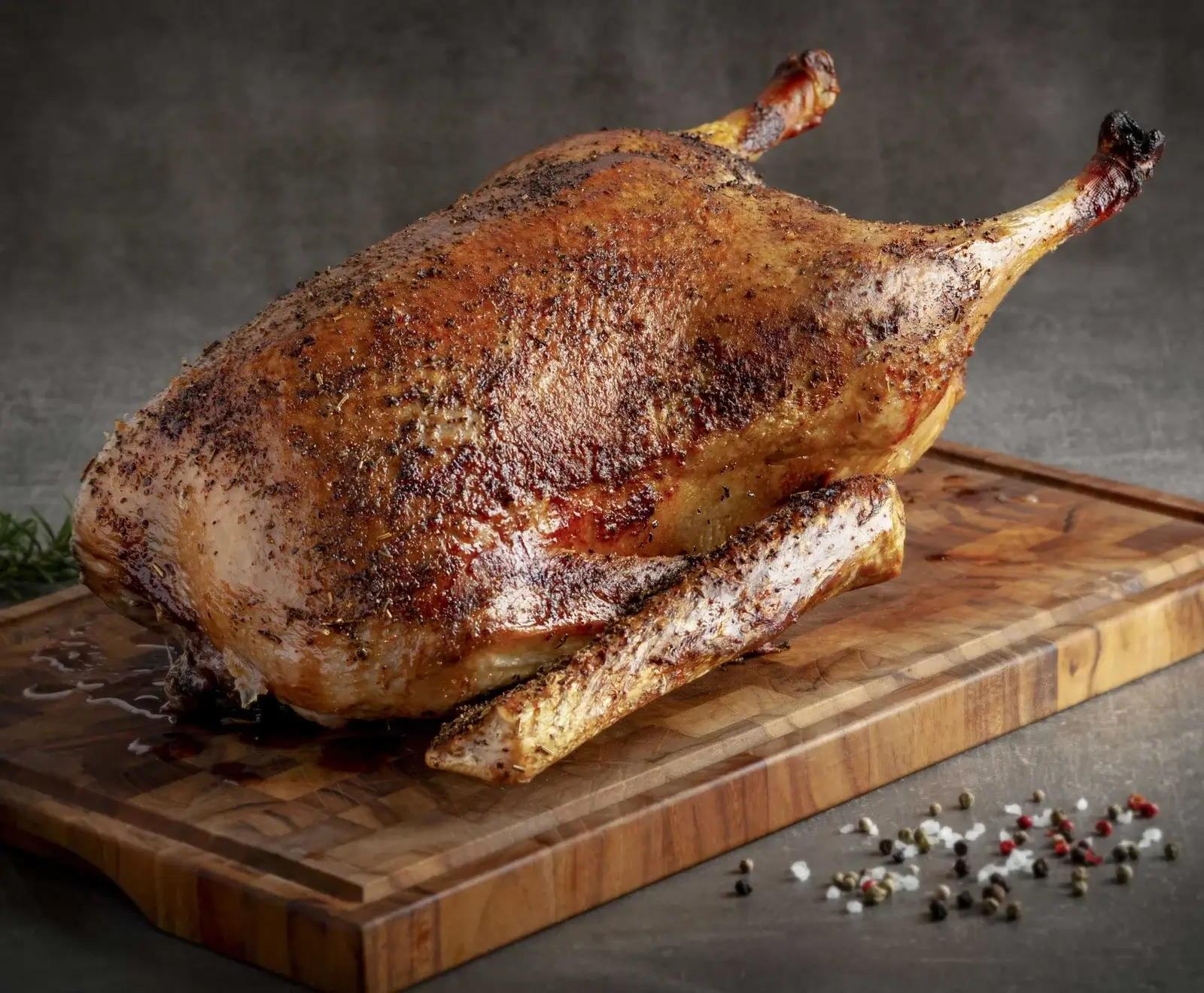 Ganze, knusprig gebratene Ente mit goldbrauner Haut und Kräutern auf einem Holzschneidebrett, umgeben von Pfefferkörnern und grobem Salz.